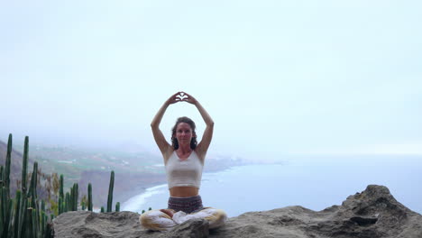 Seated-atop-a-mountain,-a-woman-meditates-and-forms-the-Maha-Sakal-hand-gesture,-framed-by-the-ocean-and-lush-green-mountains