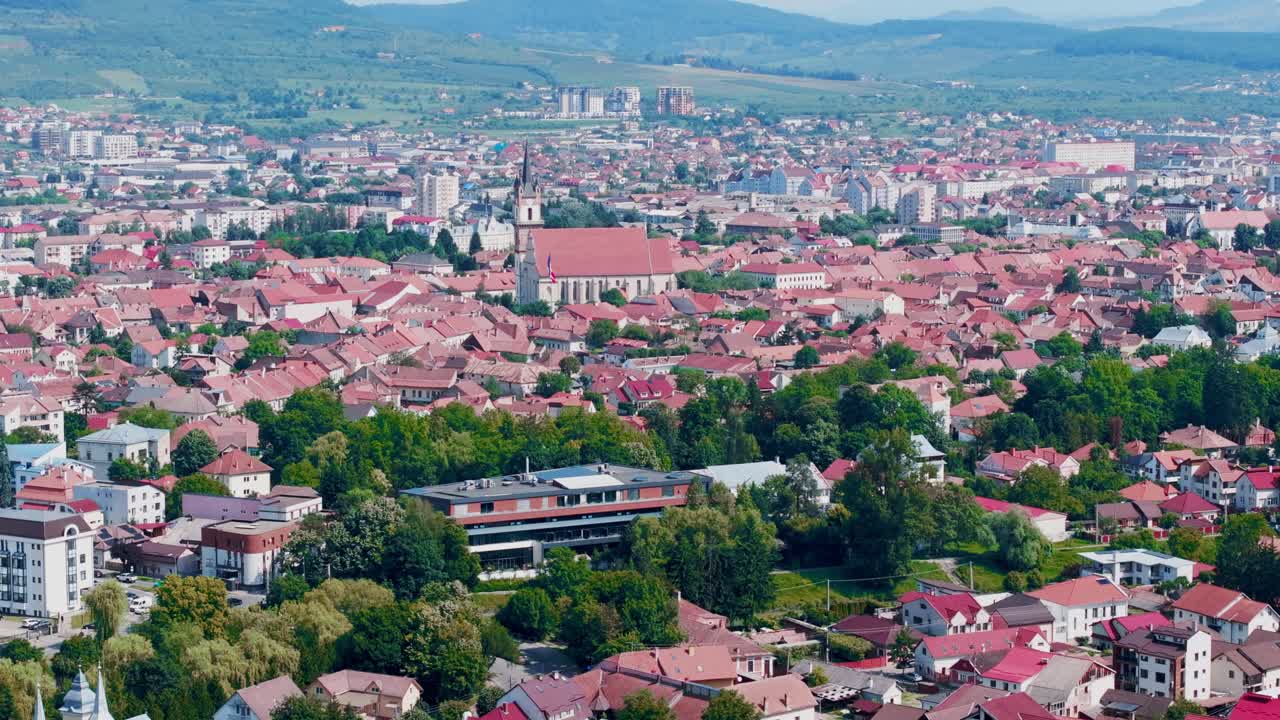 A static, wide-angle aerial photograph captures a sprawling European cityscape dominated by red-roofed residential buildings. In the background, the Evangelical Church of Bistrita
