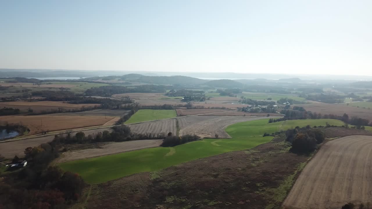 Drone footage of a patchwork rural farmland featuring green and brown crop fields, with distant hills and a lake visible along the horizon under clear skies.