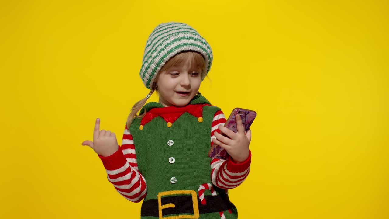 Holčička v kostýmu vánočního skřítka uskutečňuje videohovor na mobilním telefonu.