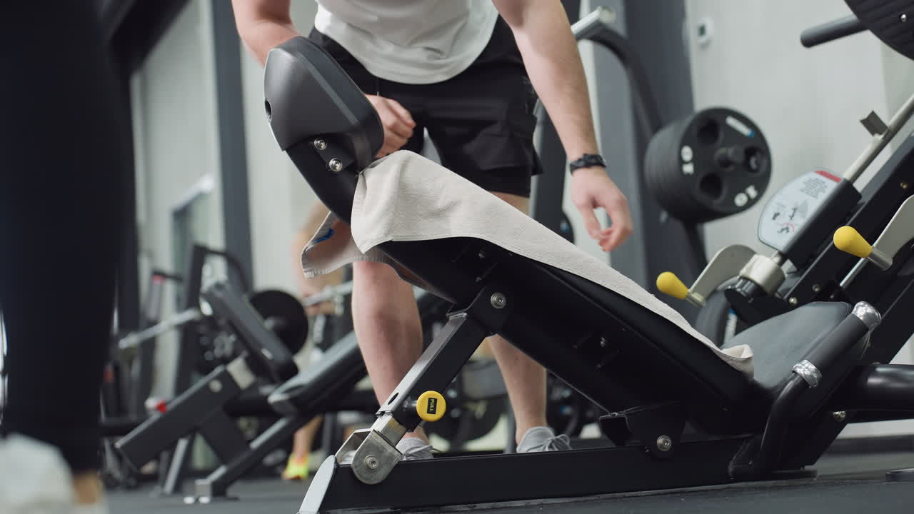 Keep fit buff spreads sports towel on weight bench then seats comfortably on machine preparing for leg press with controlled posture under modern gym lighting capturing post workout readiness focus