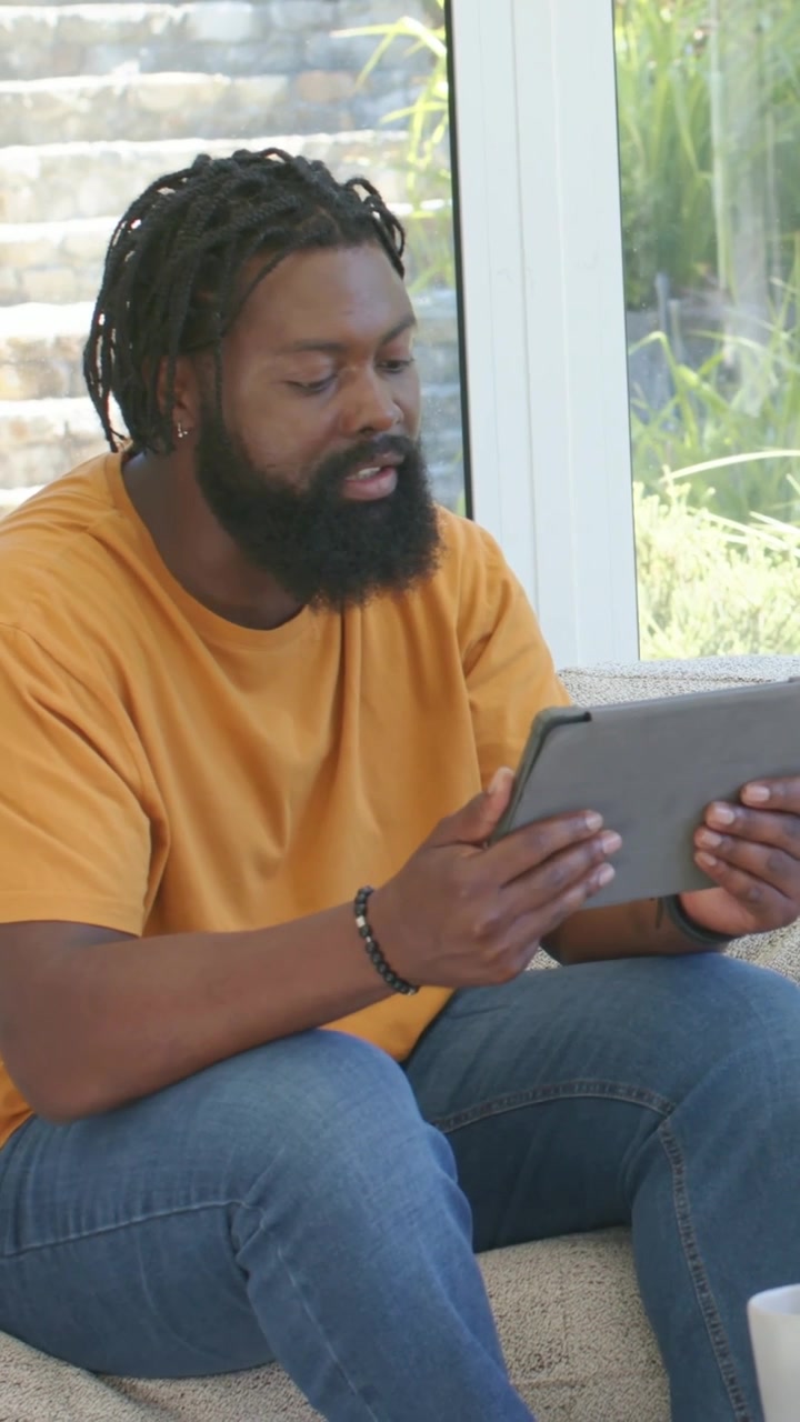 Vertical video: Tapping tablet with left hand, man with beaded bracelet browsing on couch at home
