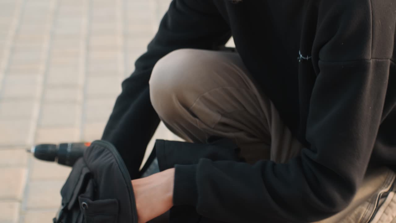 Ung mand tjekker lille værktøj og batteri. Mand knæler på fortovet og inspicerer værktøj med en skruetrækker. Ung mand hukker sig ned på byens fortov og inspicerer omhyggeligt udstyr med værktøj og batteri.