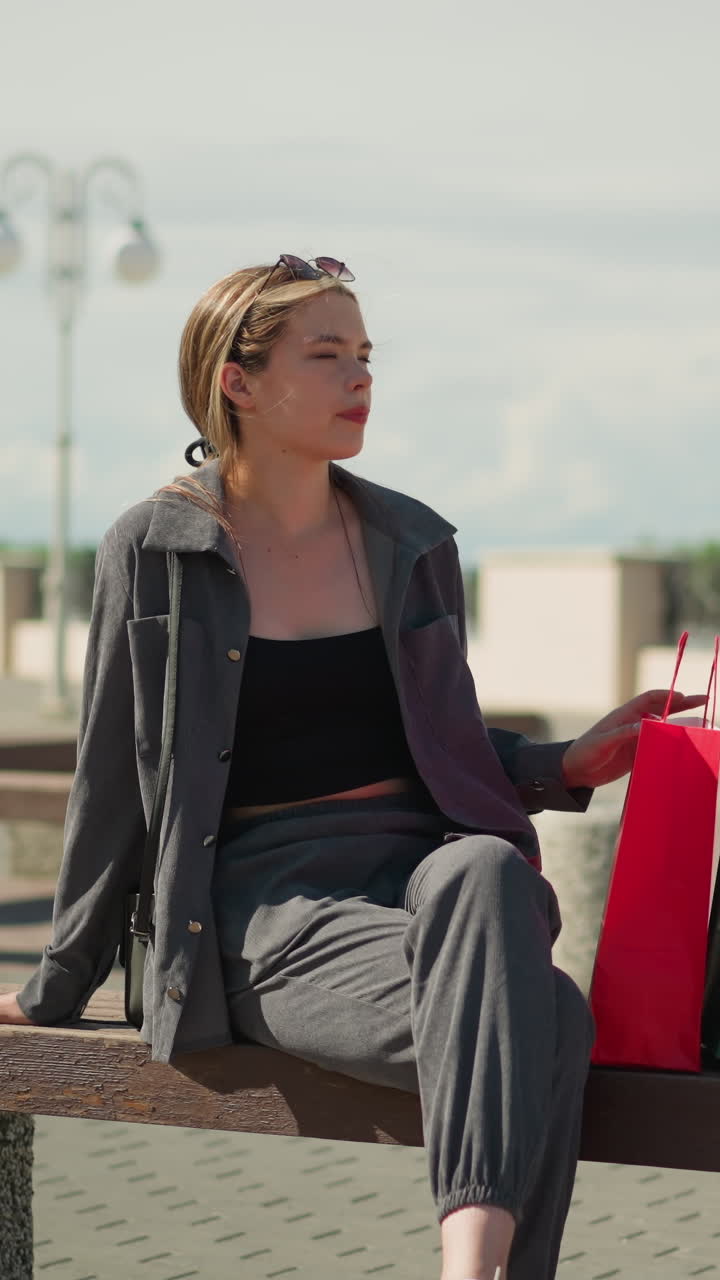 la chica blanca deja caer sus bolsas de compras en un banco, se sienta al sol, y mira a la distancia con una expresión relajada, cruza las piernas y coloca su mano derecha en su pierna