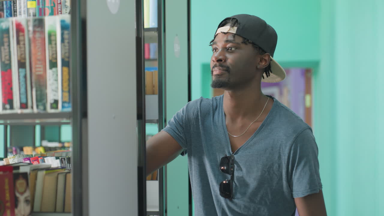 Young student wearing cap holds book while returning it to shelf inside modern library with teal walls, focused expression, and casual style