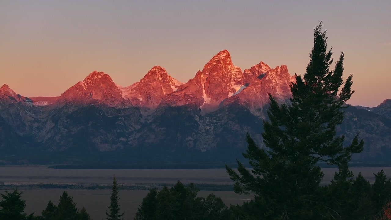 Golden sunrise alpenglow light on snowy Grand Teton mountain range above valley, sunset golden hour warmth