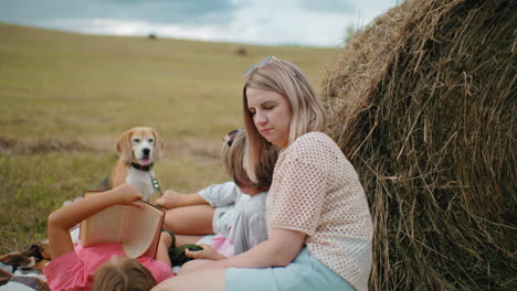 thoughtful woman with sunglasses on head laughs with friend nearby, little girl lies reading, and dog stands in background near hay bale in open countryside