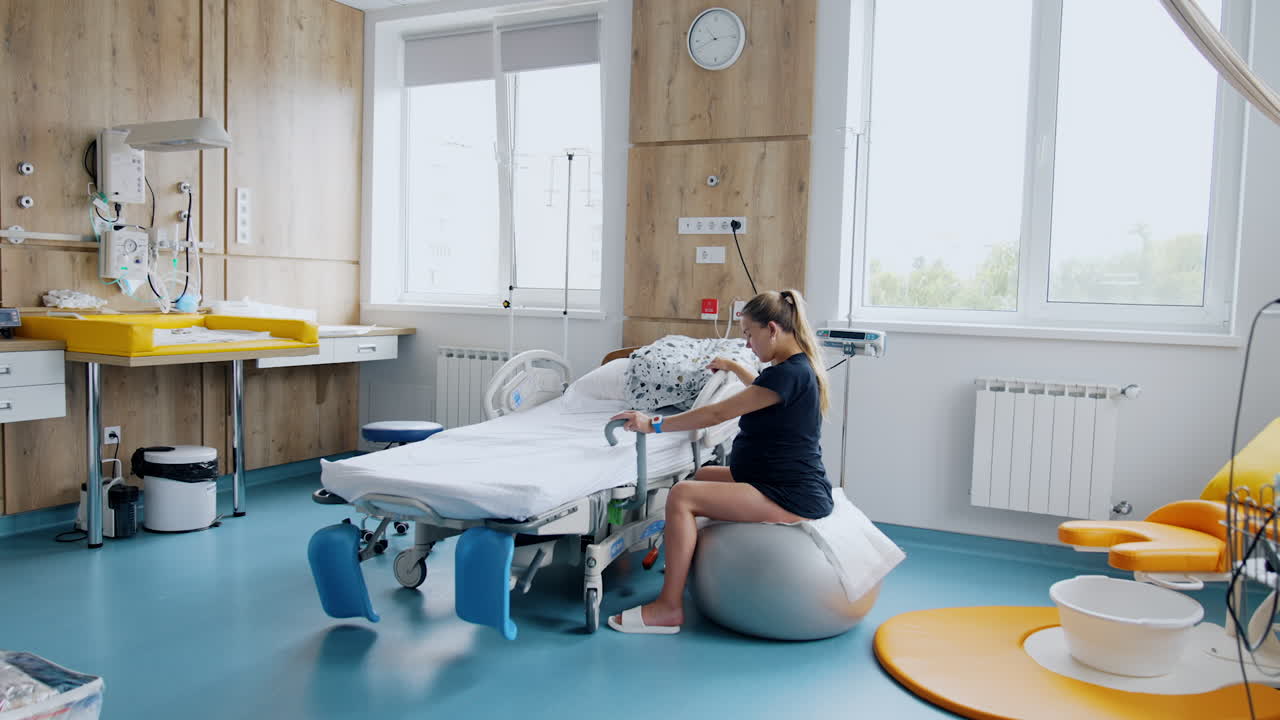 Woman expecting a baby is in the modern hospital ward. Lady sits on the fitball swaying slowly during contractions.