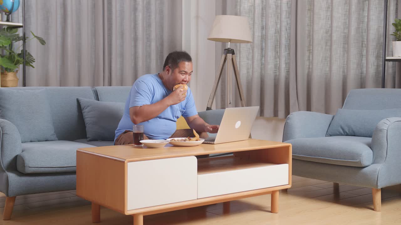 Full Body Of A Fat Asian Man Eating Fast Food Having Hamburger While Working On A Laptop In The Living Room At Home
