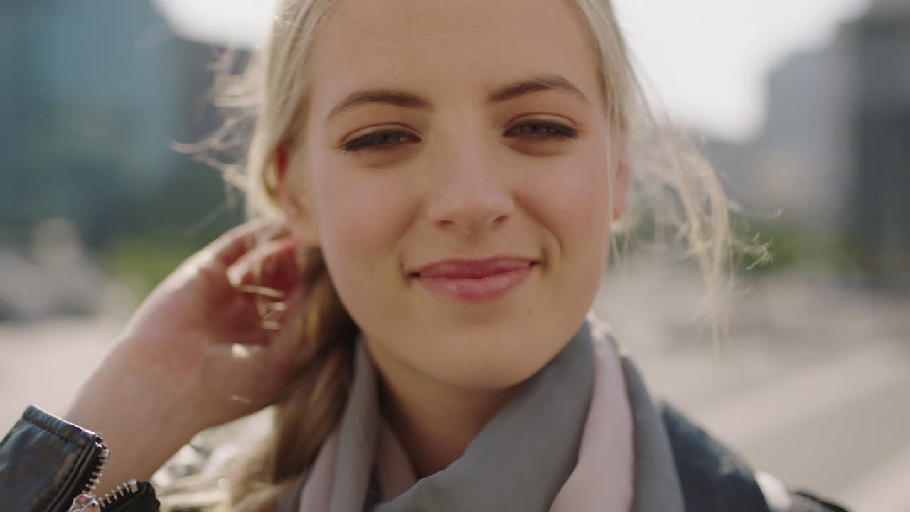 행복 한 젊은 금발 여자 학생 의 포트레이트 는 카메라 에 미소 짓고  ⁇ 빛 이 있는 도시 배경 에서 머리카락 을 통해 손 을  ⁇ 고 달리고 있다