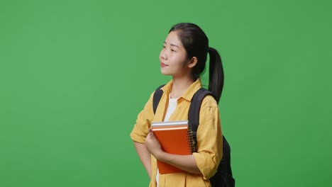 side view of asian woman student with a backpack and a book standing with one arm akimbo and looking around in the green screen background studio