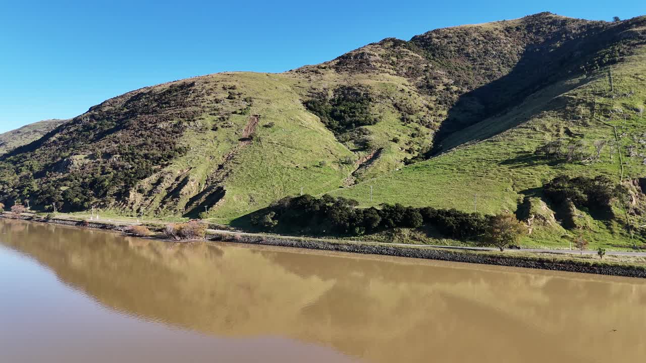Drone footage captures Akaroa's lush hills and reflective lake under clear skies, showcasing natural beauty and tranquility