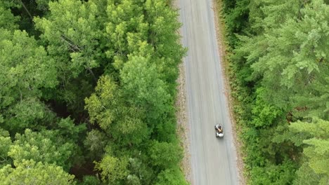 drone tracking shot going upwards of someone driving an atv on a dirt road surrounded by green pine trees in canada