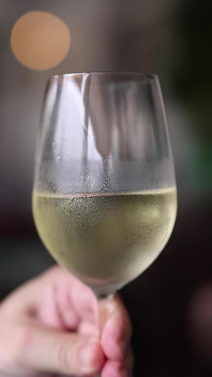 Una mano presenta un bicchiere di vino bianco coperto di condensa su uno sfondo scuro con effetto bokeh.