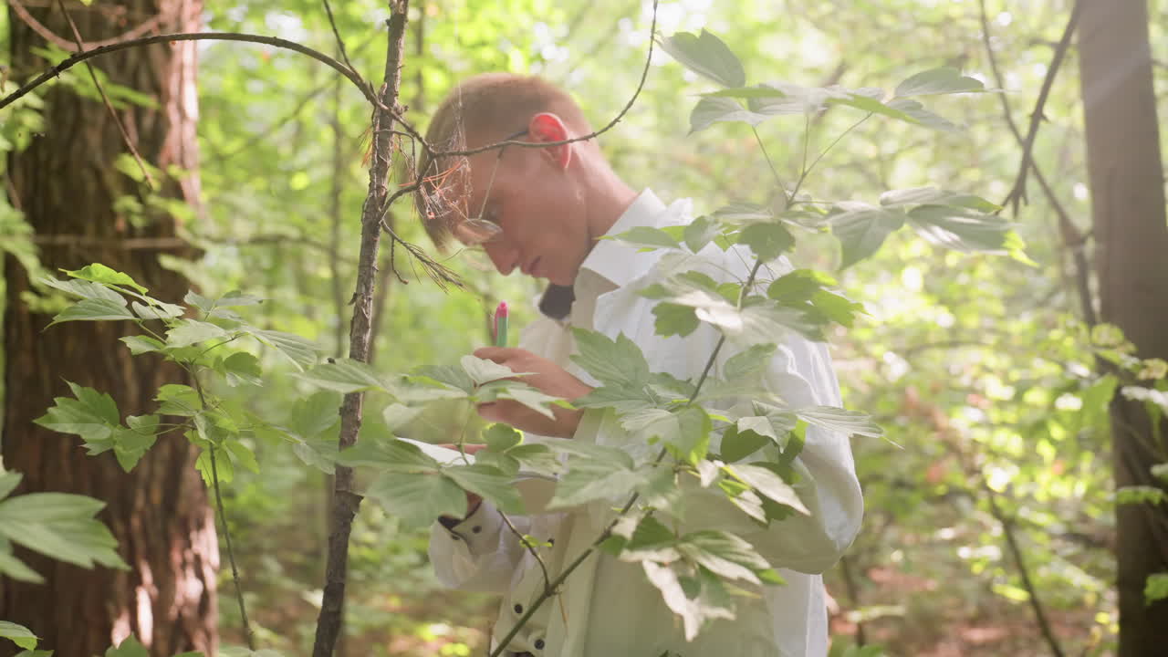 Blurred light view of botany student in white coat standing in forest under bright sunlight with serious expression writing notes in jotter using pen, documenting research among lush leaves and trees