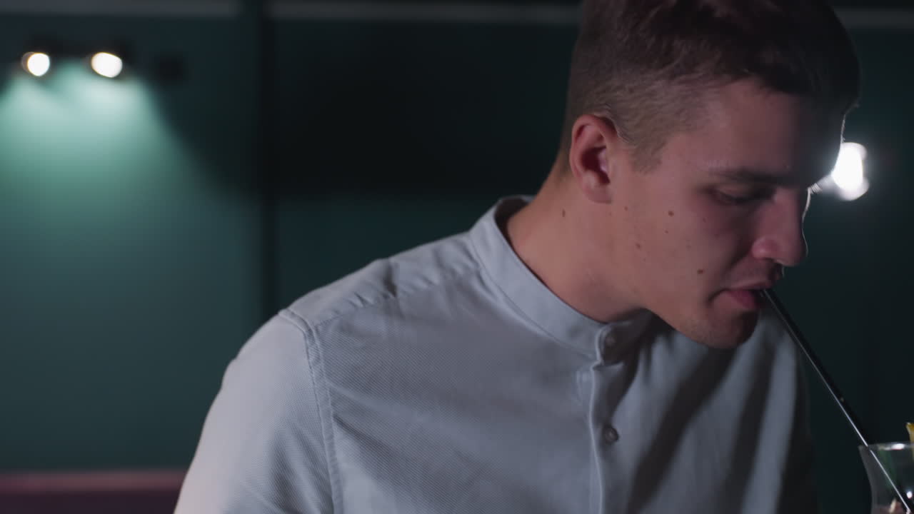 Close-up of male wearing white shirt and grey trousers, holding cue stick and glass of lemon drink with black straw. Ice cubes and lemon slice float in glass as he raises it up for a sip