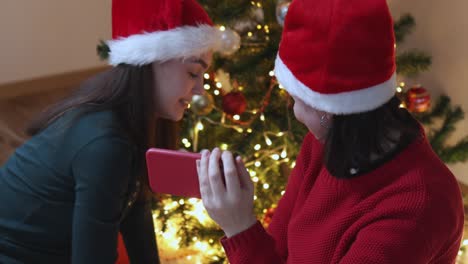 two females in green santa hat using smartphone on christmas tree background