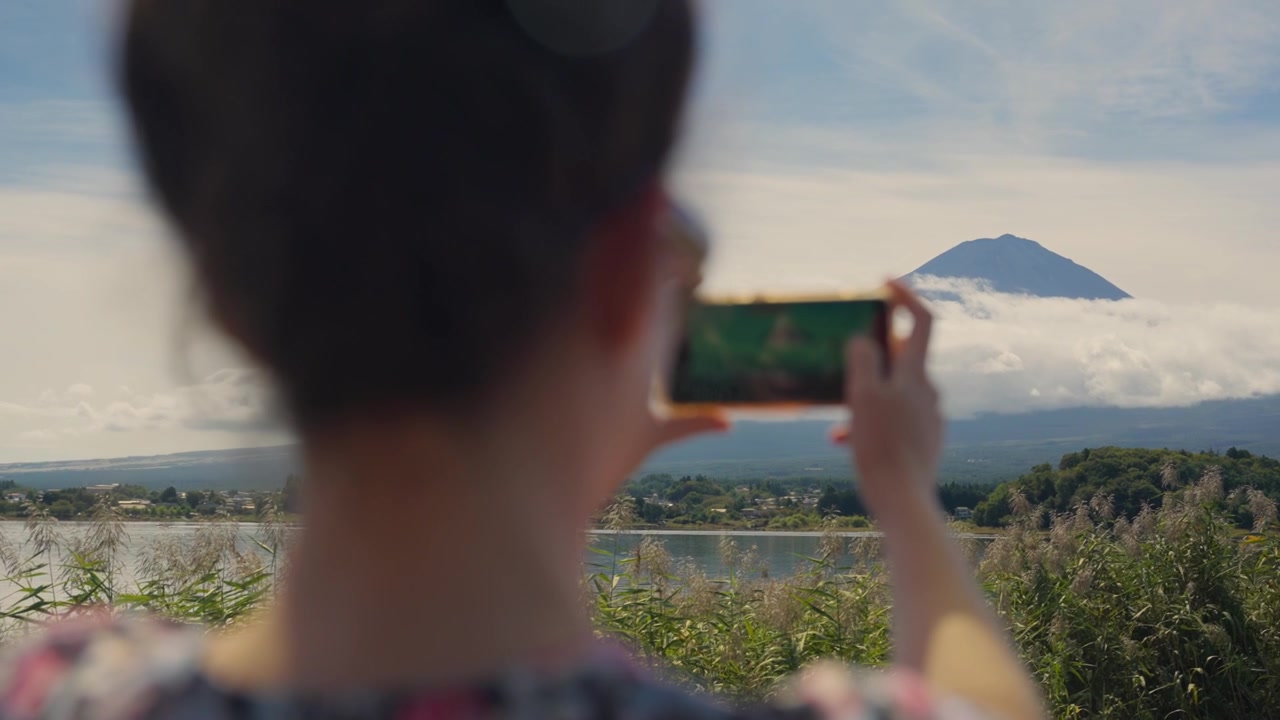 Over the shoulder view of a female tourist taking a picture of the iconic Mount Fuji in Japan with her smartphone, capturing the serene landscape and sharing her travel experience online