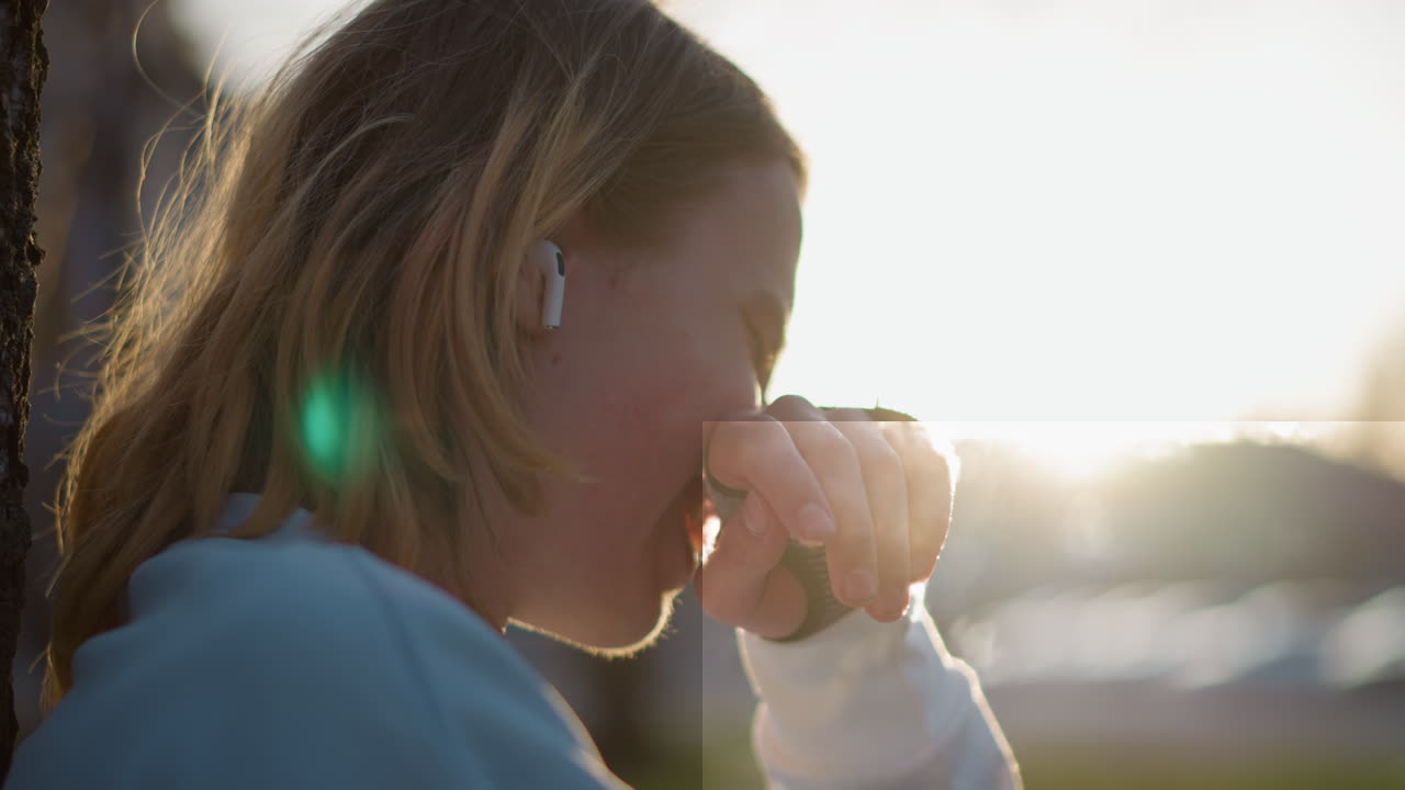 Adolescente blanco escuchando música al aire libre, bebiendo café y mirando el móvil durante una hora dorada y cálida; perfil natural, con auriculares, destellos de lente suaves y una sonrisa relajada, momento íntimo y tranquilo