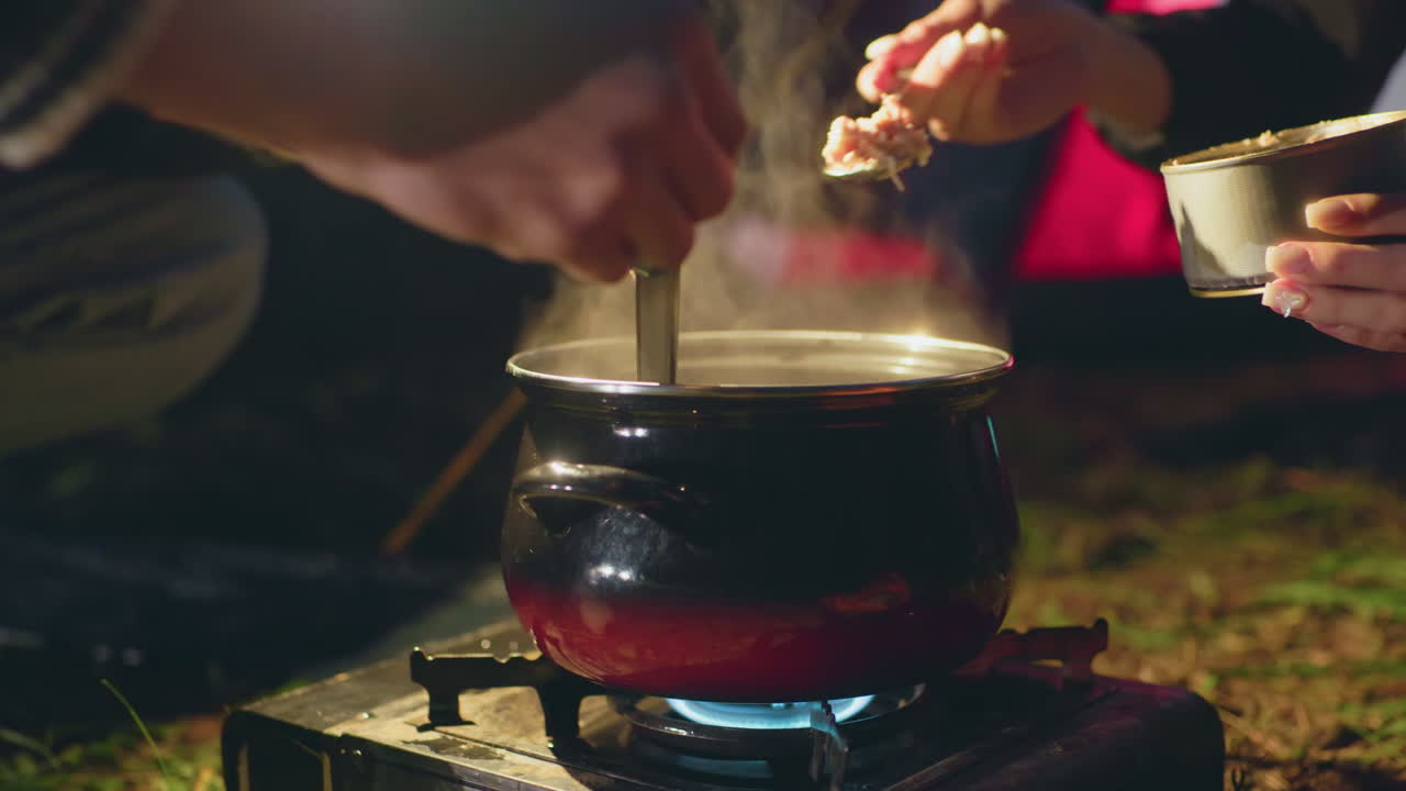Close up of two people cooking outdoors at night as one stirs boiling pot on gas burner while other adds canned food, with steam rising and warm glow from nearby tent lighting the scene