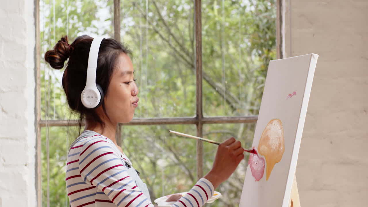 asian young woman painting on canvas while listening to music at home