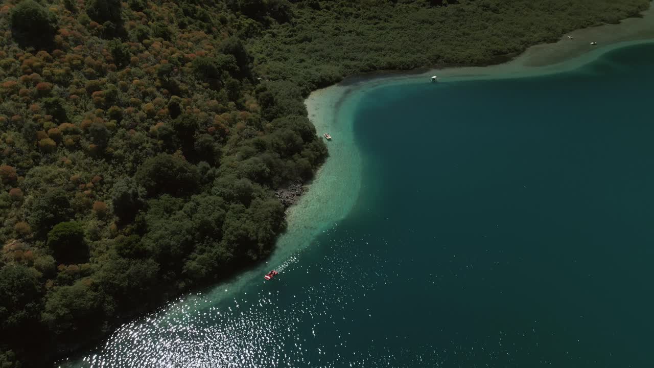 Curved shoreline of Lake Kournas with brilliant turquoise shallows contrasts with lush hills covered in greenery and spotted with shrubs in Crete, Greece