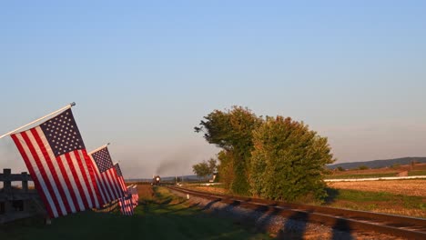 a view of a line of gently waving american flag on a fence by farmlands as a steam passenger train approaches in late afternoon