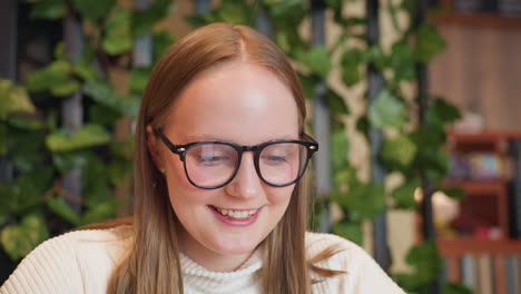 young student wearing glasses and white sweater smiles while seated in cozy cafe with blurred background featuring warm lighting, leafy wall decor and striped counter