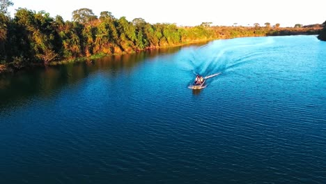 A-small-fishing-boat-travels-down-a-deep-blue-river-in-the-evening-sun,-SLOW-MOTION-aerial-drone-shot