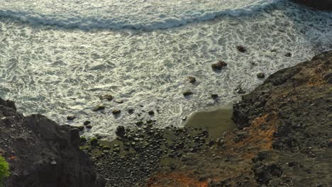Waves-roll-on-secluded-rocky-beach-with-bay-cliffs-in-evening-sunlight