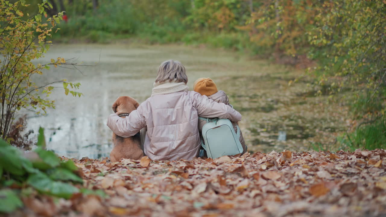 woman places arm around dog and child as they sit by calm leaf strewn lake in autumn forest sharing warm affectionate family bond amid tranquil natural setting