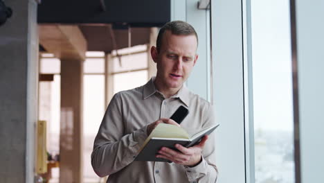 young man leaned on the window holding a telephone and a paper notebook. man turning pages in a book and comments something.