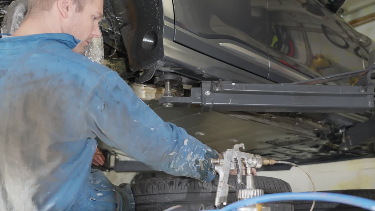 Man sitting beneath car on lift removes cap, inserts spray gun tube into sill cavity, applying lanolin rustproofing