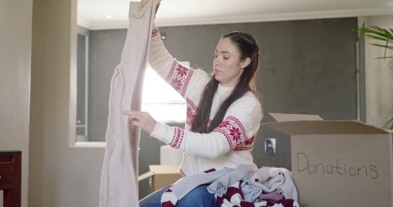 Woman pulling gray top from pile at home, checking, folding, placing into Donations box donating
