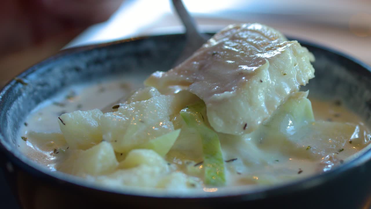 Hand lifts creamy haddock, potatoes, and leeks from bowl in natural daylight, shallow focus