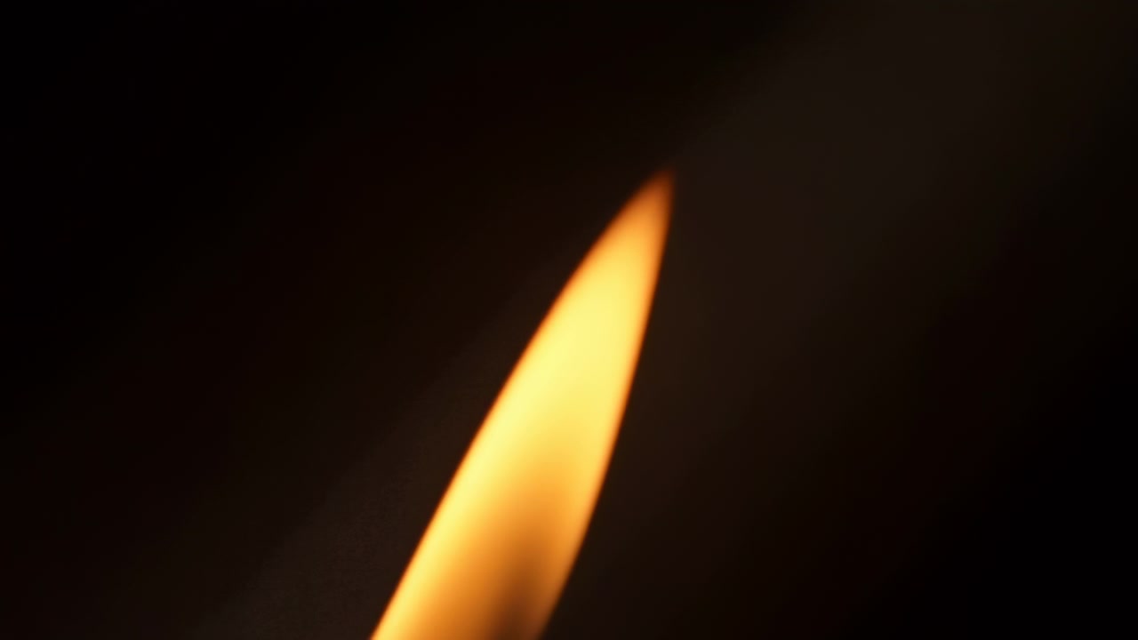 Intimate close-up of single candle flame with elongated narrow tip angled slightly right, steady luminous burn, smooth yellow to amber gradient, dark isolated background, calm mood