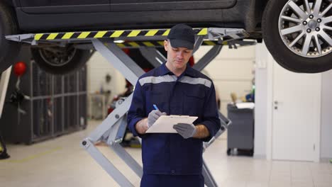 A-car-mechanic-standing-next-to-lifted-car,-making-notes-on-a-tablet