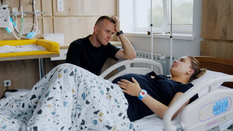 man sits above his wife lying in maternity hospital bed. woman strokes her pregnant belly saying something with her eyes closed.