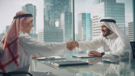 dos empresarios emiratíes felices en kandura tradicional blanca sentados en la oficina y firmando el contrato. los socios comerciales se estrechan la mano en una inversión lucrativa. el concepto de empresario saudí, emiratí y árabe.