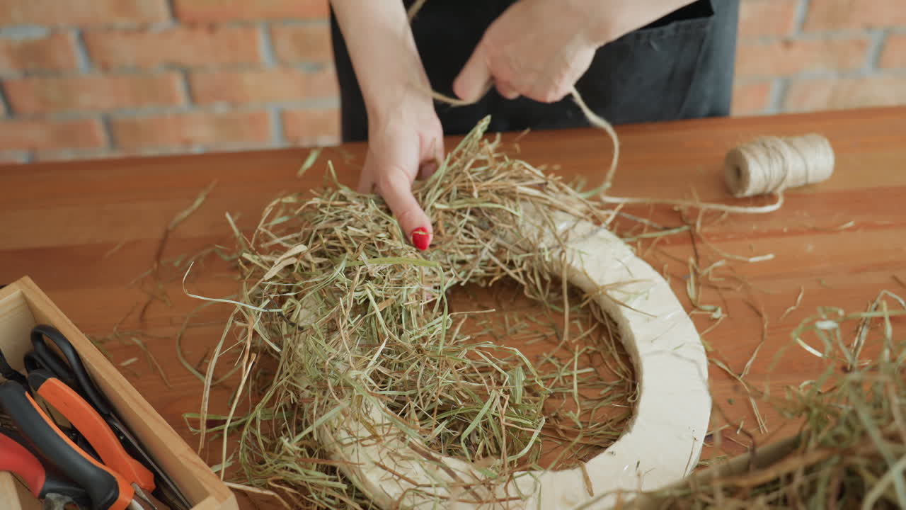 Decorator arranging dried hay on circular base with twine spool and tools nearby, preparing rustic wreath craft project on wooden table in creative workshop focused on handmade seasonal decoration design