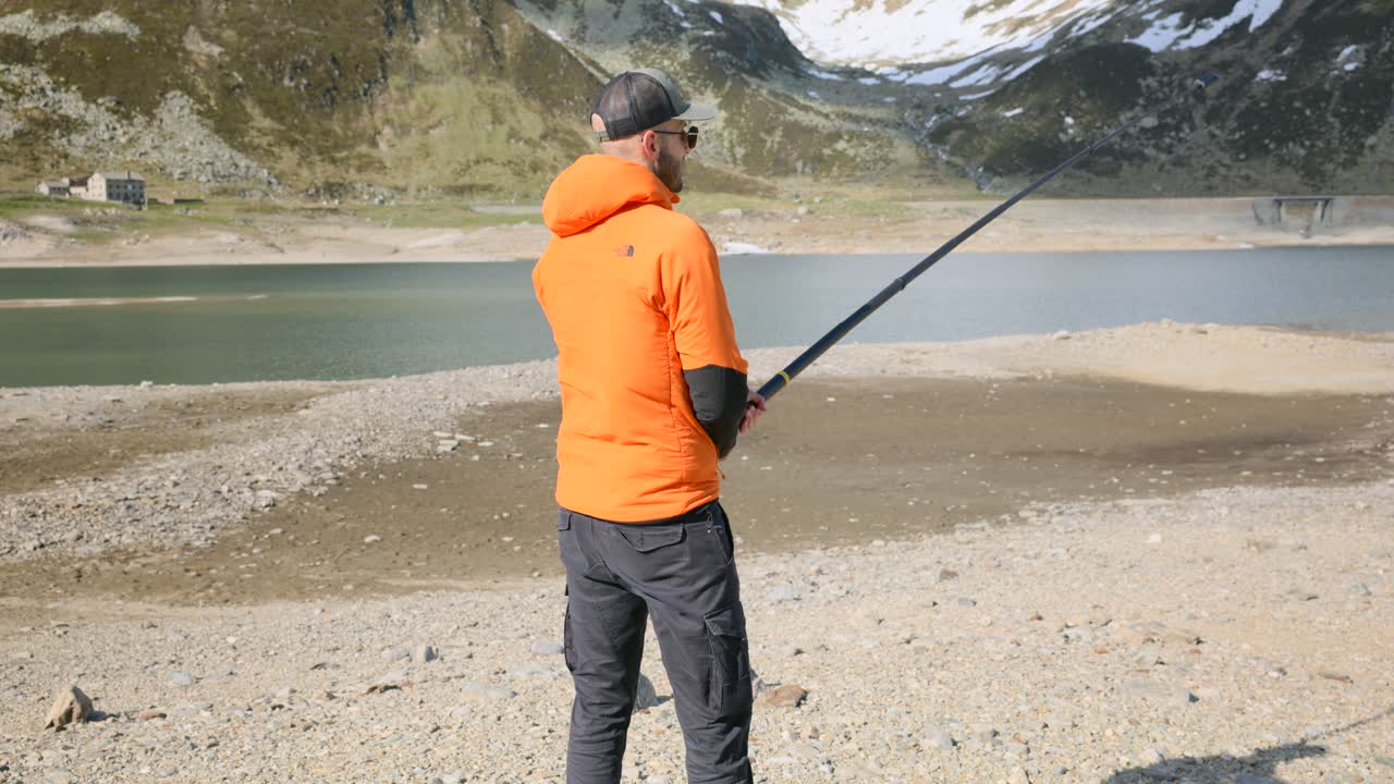 Man in orange jacket films scenic mountain lake with 360 camera on a long stick, capturing immersive outdoor footage in a remote, high-altitude landscape