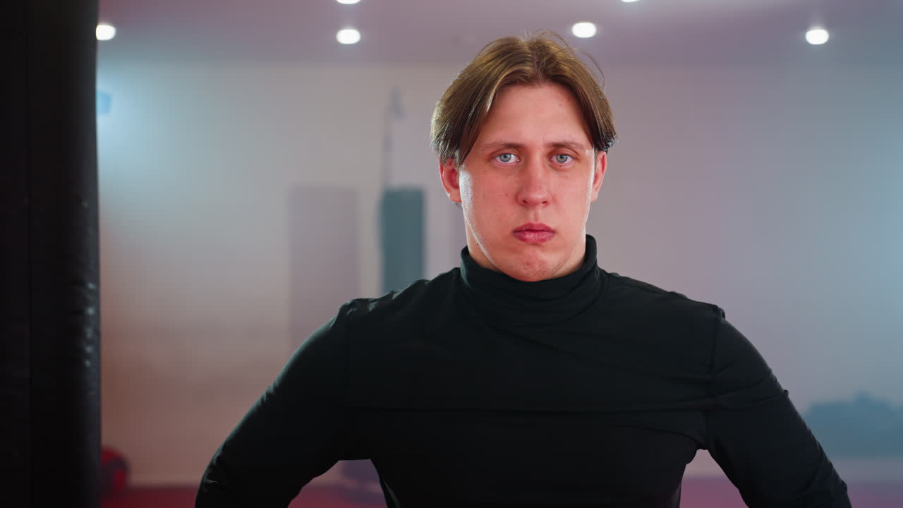Male in black long sleeve shirt standing in gym near punching bag, looking to side with serious focused expression, illuminated by bright lights, representing training, discipline, strength, confidence