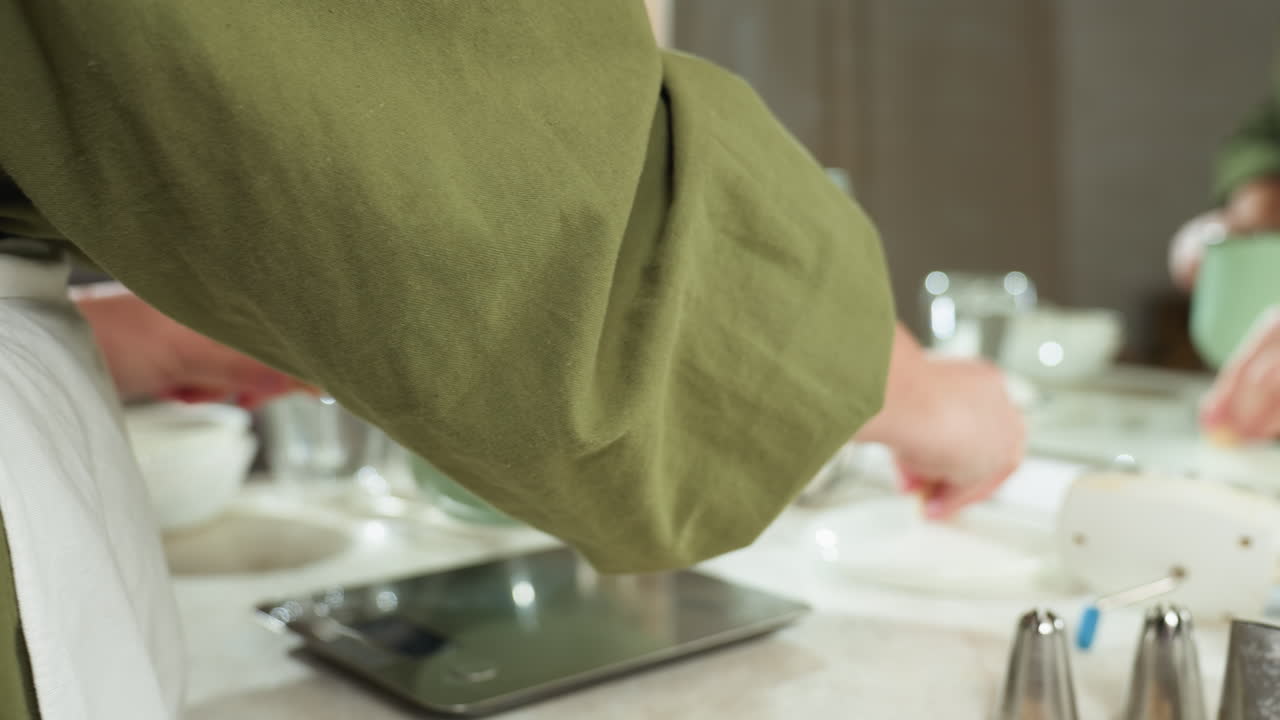Close up of woman in green sleeve removing pot lid and placing pot on electric stove, turning it on, then reaching for ingredients from basket to add, surrounded by kitchen items and digital scale