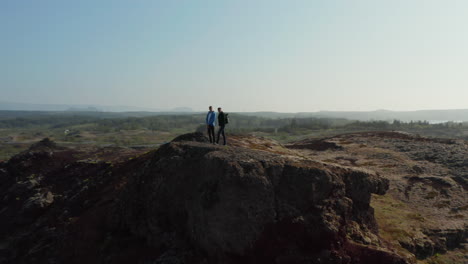 Drohnenansicht-Von-Zwei-Männern,-Die-über-Einer-Klippe-Stehen-Und-Ein-Panorama-In-Island-Betrachten.-Drohnenansicht-Von-Zwei-Rucksacktouristen,-Die-Die-Landschaft-Von-Island-Nach-Dem-Erklimmen-Eines-Hügels-Genießen