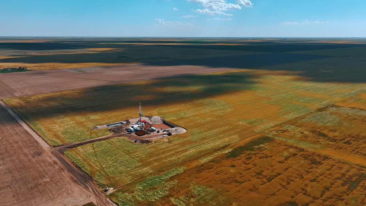 Flying closer to the little location in the dessert for drilling the natural resources. Clouds throw shadows on the immense field on sunny day.