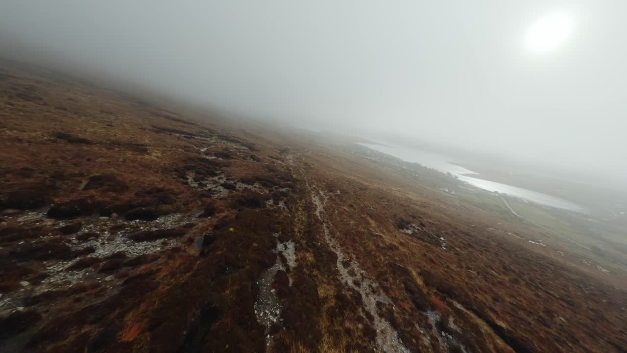 FPV drone flying fast down a hill covered by low hanging clouds towards a small village on Achill Island
