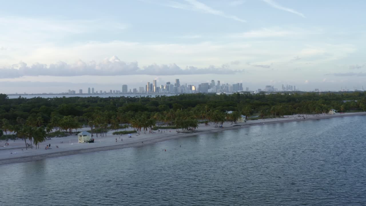dolly en la inclinación aérea de la playa tropical rodeada de palmeras en el parque crandon en key biscayne con el horizonte de miami, florida en la distancia en una soleada noche de verano