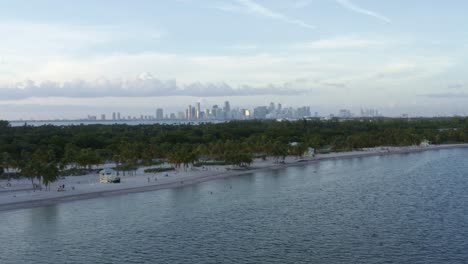 dolly in tilting up aerial drone shot of the tropical beach surrounded by palm trees on crandon park in key biscayne with the skyline of miami, florida in the distance on a sunny summer evening