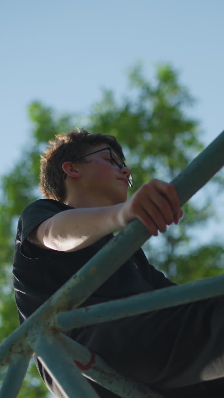 어린 소년은 야외의 파란 철 구조물 위에 서 있으며, 두 손으로 프레임을 잡고 사려 깊게 보이며, 은 축구 공이 철 막대 안에 혀 있습니다.