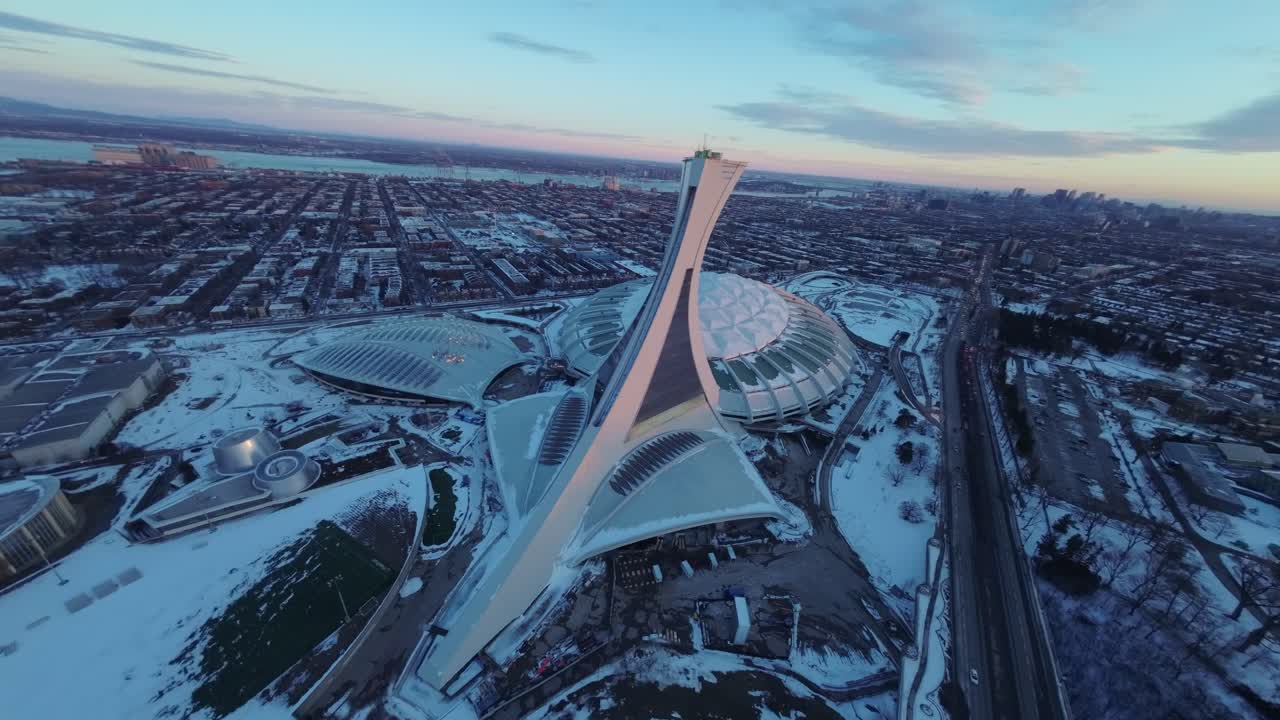 FPV drone captures sweeping aerial view of Olympic Stadium in Montreal at dusk, snow-dusted winter scene